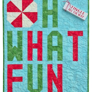 Peut inclure: Une tenture murale matelassée avec les mots "OH WHAT FUN" en lettres majuscules rouges et vertes sur un fond bleu clair. Un motif géométrique rouge et blanc est dans le coin supérieur gauche. Une petite étiquette rectangulaire indique "JINGLE ALL THE WAY".