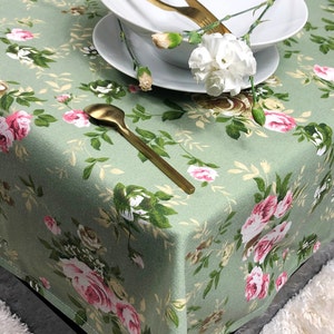 Könnte beinhalten: Eine grüne Tischdecke mit einem Blumenmuster mit rosa und weißen Rosen. Die Tischdecke ist aus gewebtem Stoff und hat eine dezente Textur. Ein goldener Löffel und ein weißer Teller mit einer weißen Nelke liegen auf der Tischdecke.