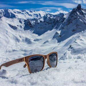 Peut inclure: Lunettes de soleil en bois avec des verres fonc&eacute;s reposant sur un sommet de montagne enneig&eacute;. Les lunettes ont une monture en bois brun et le nom de la marque "Sunfox" est visible sur la branche droite.