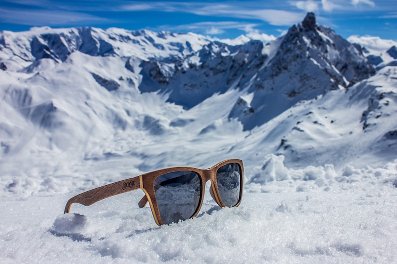 Peut inclure: Lunettes de soleil en bois avec des verres fonc&eacute;s reposant sur un sommet de montagne enneig&eacute;. Les lunettes ont une monture en bois brun et le nom de la marque "Sunfox" est visible sur la branche droite.