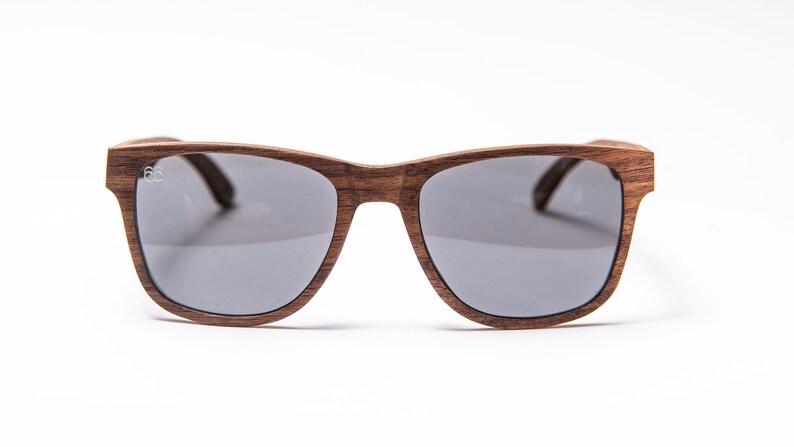 Peut inclure: Une paire de lunettes de soleil en bois brun avec des verres gris. Les lunettes ont une forme rectangulaire simple.