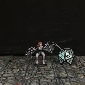May include: A miniature figurine of a winged humanoid figure with red hair and a dark outfit, posed next to a large, twenty-sided die. The die has a teal glow and white numbers. The background is dark.