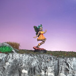 May include: A miniature fairy figurine with a yellow and orange dress, green wings, and teal hair. The fairy is standing on a red base, set against a purple and pink sky. The figurine is on a rocky cliff with green grass.