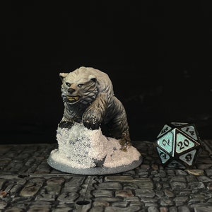 Puede incluir: Una figura de oso en miniatura pintada a mano, en tonos grises y blancos, posada sobre una base nevada. Junto a ella, un dado de veinte caras con acabado turquesa y negro, ideal para juegos de mesa.