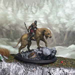 May include: A miniature figurine of a warrior riding a large feline creature. The warrior is clad in armor and holds a spear. The feline is tan with a snarling expression, mounted on a rocky base. The background shows a blurred forest.