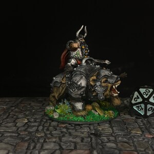 Puede incluir: Figura en miniatura de un guerrero montado en un oso, ambos con armadura. El guerrero tiene un casco con cuernos y una capa roja. Un dado de veinte caras en primer plano, tema de juego de mesa.