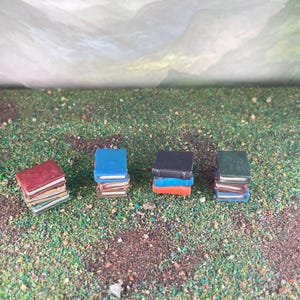 May include: Four stacks of miniature books with red, blue, black, and green covers. The books are on a textured green surface with a blurred background. Each stack has a different number of books.