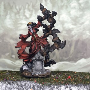 Puede incluir: Figura en miniatura de una mujer vampiro con un vestido rojo, rodeada de murciélagos. La figura se encuentra sobre una base de lápida. El fondo muestra un paisaje borroso.