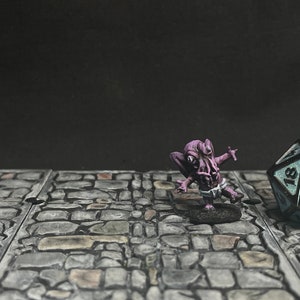 Puede incluir: Una figura en miniatura de un monstruo morado con tentáculos y un taparrabos se encuentra sobre una base de piedra. Un dado grande de múltiples caras con números blancos está cerca, sobre una superficie de piedra, sugiriendo un juego de mesa.