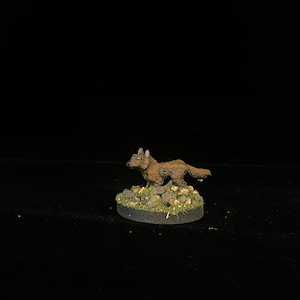 Puede incluir: Figura en miniatura de un lobo marrón en posición de carrera. El lobo está de pie sobre una base de rocas y vegetación verde. La figura es probablemente parte de un juego de mesa o exhibición.