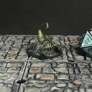 Puede incluir: Una figura de escorpión en miniatura con un acabado verde metálico y negro, colocada sobre una base rocosa. Un dado de veinte caras con detalles turquesa está cerca. La escena está ambientada sobre una superficie pavimentada con piedras, lo que sugiere un juego de mesa.