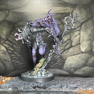 Puede incluir: Figura de fantasía en miniatura de una criatura con rostro esquelético, armadura y alas moradas. La figura está montada sobre una base redonda y se alza sobre una superficie rocosa. Una estructura arbórea se extiende desde la base.
