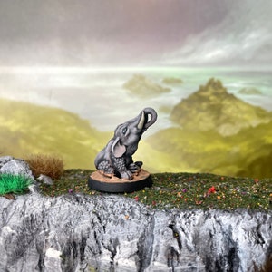 Puede incluir: Figura de elefante gris con la trompa levantada, sentado sobre una base redonda y oscura. El elefante tiene piel texturizada y colmillos. El fondo presenta un paisaje con cielo nublado y colinas.