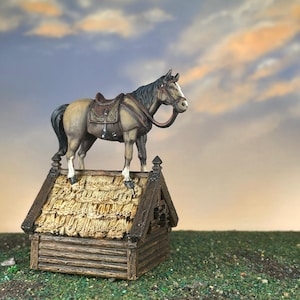 May include: A miniature figurine of a horse with a saddle and bridle stands atop a small wooden structure with a thatched roof. The horse is tan with a black mane and tail. The structure is brown and appears to be a miniature well.
