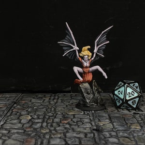 May include: Miniature figurine of a winged woman with blonde hair and an orange dress, perched on a rock. The figure has large bat-like wings. A glowing, multi-sided die is in the foreground. The background is dark.
