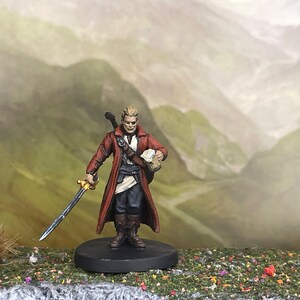May include: Miniature figure of a character in a red coat, holding a sword. The figure has blonde hair and is standing on a black base. The background features a painted landscape of rolling hills.