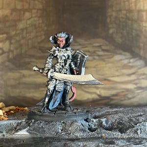 May include: Miniature figurine of a fantasy character in silver armor, holding a large sword and shield. The figure has horns and a tail, standing on a stone base. The background is a blurred, textured wall.