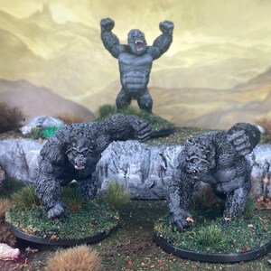 Könnte beinhalten: Drei graue Gorilla-Figuren auf einer grasbewachsenen Basis, eine steht mit erhobenen Armen, zwei weitere in geduckter Haltung. Der Hintergrund zeigt eine Landschaft mit Felsen und einem gedämpften Himmel.
