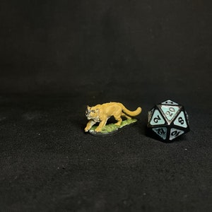 Puede incluir: Una figura en miniatura de un puma en posición de caminar, pintada en tonos beige y marrón, se encuentra sobre un parche verde. Junto a ella hay un dado negro y verde azulado de veinte caras, usado en juegos de rol.
