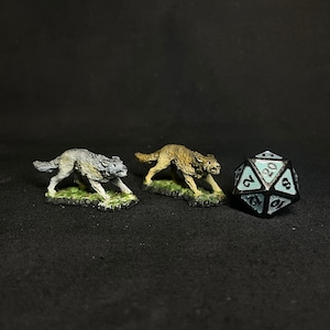 Puede incluir: Dos figuras de lobos en miniatura, una gris y otra marrón, sobre bases verdes. Un dado de veinte caras con detalles en negro y verde azulado está cerca. La escena se desarrolla sobre un fondo oscuro.