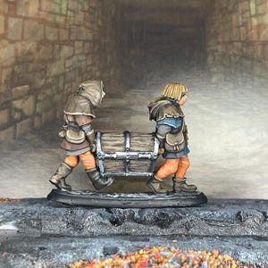 Puede incluir: Figuras en miniatura de dos personas que transportan un cofre de madera. Las figuras visten ropa de estilo medieval, con capas marrones y pantalones naranjas. El cofre está detallado con bandas y cierres metálicos. La escena está ambientada en un túnel de piedra.