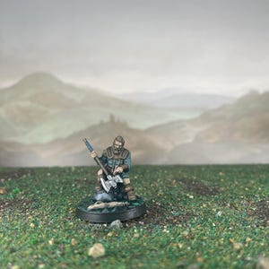 May include: Miniature figurine of a bearded man in a teal and brown outfit, holding a silver axe. The figure is kneeling on a circular base with a small fire pit, set against a backdrop of rolling hills and a cloudy sky. The miniature is likely for tabletop gaming.