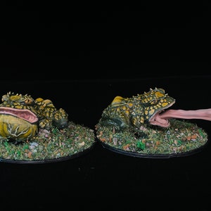 Puede incluir: Dos figuras de ranas en miniatura detalladas. Una rana está con la boca cerrada, la otra tiene la boca abierta con una larga lengua extendida. Ambas están pintadas en tonos verdes, amarillos y marrones, y están sobre una base de hierba artificial y pequeñas rocas.