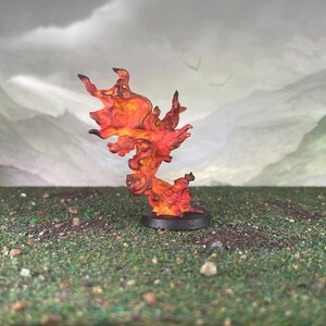 May include: A miniature, stylized flame sculpture. The sculpture is primarily orange and red, with darker accents. It is mounted on a small, round, black base. The background features a landscape with a cloudy sky and green hills.