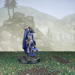 May include: A miniature figurine of a female figure with a skull-like face, draped in a flowing blue cape. The figure is painted in shades of gray, blue, and red, and is mounted on a circular base. The background features a blurred landscape.