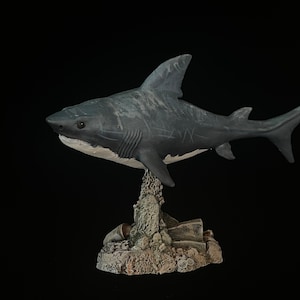 Puede incluir: Figura de tiburón gris y blanco con marcas de arañazos, montada sobre una base rocosa texturizada. El tiburón tiene una aleta dorsal detallada sobre un fondo negro. Artículo decorativo.