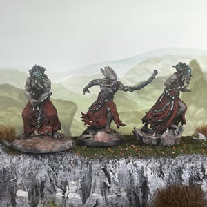 Puede incluir: Tres figuras de fantasía en miniatura con piel gris y ropas rojas rasgadas. Cada figura está posada de manera diferente, de pie sobre una base rocosa. Las figuras tienen rasgos faciales detallados sobre un fondo de paisaje borroso.