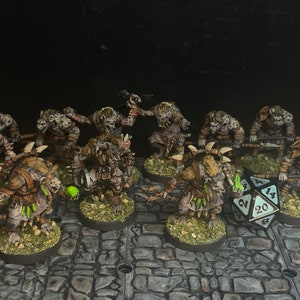 Könnte beinhalten: Eine Sammlung handbemalter Fantasy-Miniaturen, die goblinartige Kreaturen mit detaillierter Rüstung und Waffen zeigen. Die Figuren stehen auf strukturierten Sockeln, mit einem großen zwanzigseitigen Würfel. Die Farbpalette umfasst Braun-, Grün- und Grautöne.