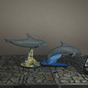 Puede incluir: Dos figuras de delfines grises, una saltando desde una base de olas azules, la otra posada sobre una estructura rocosa. Un dado de veinte caras está en primer plano, sugiriendo un tema de juego de mesa.