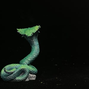 Puede incluir: Figura detallada de una cobra en una pose llamativa, con su capucha extendida y la boca abierta. La serpiente está pintada en tonos verdes y azules, con un aspecto texturizado y escamoso. La figura se encuentra sobre un fondo negro.