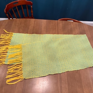 May include: Two rectangular, woven placemats with a light green and yellow pattern. The edges have a yellow fringe. The placemats are displayed on a round, wooden table. The background includes a wooden chair and a dark blue wall.