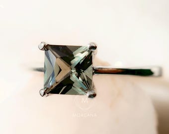 May include: A silver ring with a square-cut, pale green gemstone. The gemstone is held in place by four prongs, and the band is slender and polished. The ring is shown on a soft, off-white surface.
