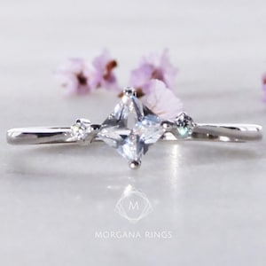 May include: A silver ring with a square-shaped clear gemstone and two smaller clear gemstones on either side. The ring is on a white surface with pink flowers in the background.