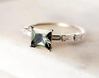 May include: A silver ring with a square, green gemstone at its centre. The band is embellished with smaller, rectangular, clear gemstones. The ring is set against a soft, white background, accentuating the jewellery's details.