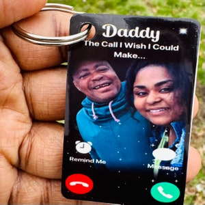 May include: Black and white phone-shaped keychain with a photo of a man and woman. The text "Daddy The Call I Wish I Could Make..." is printed on the keychain. The keychain has a "Remind Me" button and a "Message" button.
