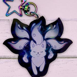 May include: A keychain featuring a white fox with nine tails, set against a dark blue and purple background. The fox is smiling, and the keychain has a star-shaped clasp with a rainbow finish.