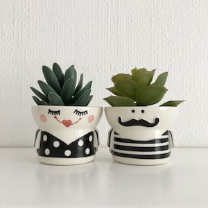 May include: Two ceramic succulent planters. One features a lady's face, a black polka dot dress, and a red heart-shaped mouth. The other has a gentleman's face, a black moustache, and a black and white striped shirt. Each planter holds a green succulent.