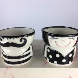 May include: Two ceramic egg cups, one with a black and white striped design and a moustache, the other with a black and white polka dot design and a smiling face.