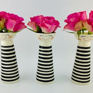 May include: Three white ceramic vases with black stripes and a smiling face design. Each vase is filled with pink roses.