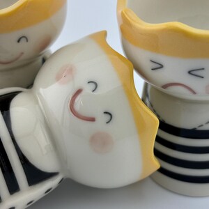 May include: Three ceramic egg cups with yellow and black stripes and a cute cartoon face. Each cup has a different facial expression.