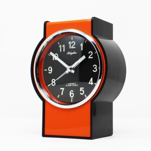 Puede incluir: Un reloj despertador Rhythm vintage con esfera negra, números blancos y manecillas plateadas. El reloj tiene una carcasa negra y naranja. El texto "Rhythm" y "2 JEWELS ALARM CLOCK" son visibles en la esfera.