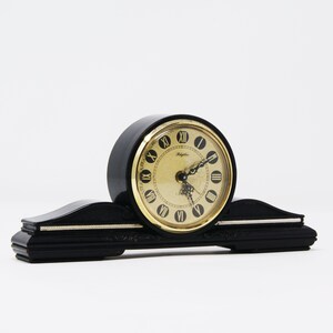 Pu&ograve; includere: Orologio da mensola nero con quadrante dorato e numeri romani. L'orologio ha una base curva e rettangolare con dettagli decorativi e un quadrante circolare. Sul quadrante &egrave; stampata la parola "Rhythm".