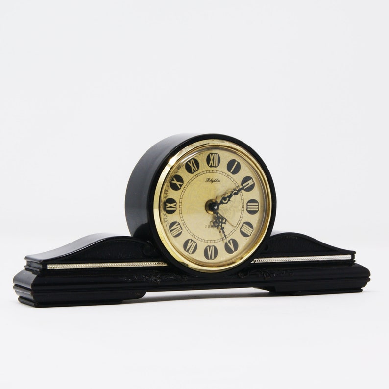 Pu&ograve; includere: Orologio da mensola nero con quadrante dorato e numeri romani. L'orologio ha una base curva e rettangolare con dettagli decorativi e un quadrante circolare. Sul quadrante &egrave; stampata la parola "Rhythm".