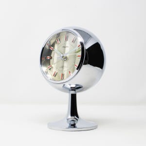 Puede incluir: Un reloj despertador esférico cromado con esfera plateada y detalles rojos y verdes. El reloj tiene la marca "Rhythm" y se apoya en una base de pedestal. Un reloj de estilo retro.