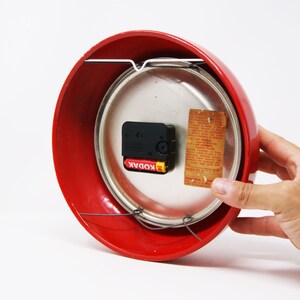 May include: Red round clock with a silver face and a black clock mechanism. The clock has a white wire hanger and a label that says "Kodak" and "Garanzia 1 anno".