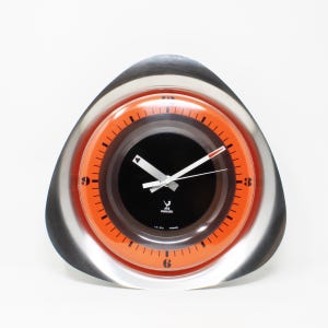 May include: A silver and orange clock with a black face and white hands. The clock is shaped like a triangle and has the text "JAZ  HORLOGERIE  LIC.  ART.  FRANCE" on the face.